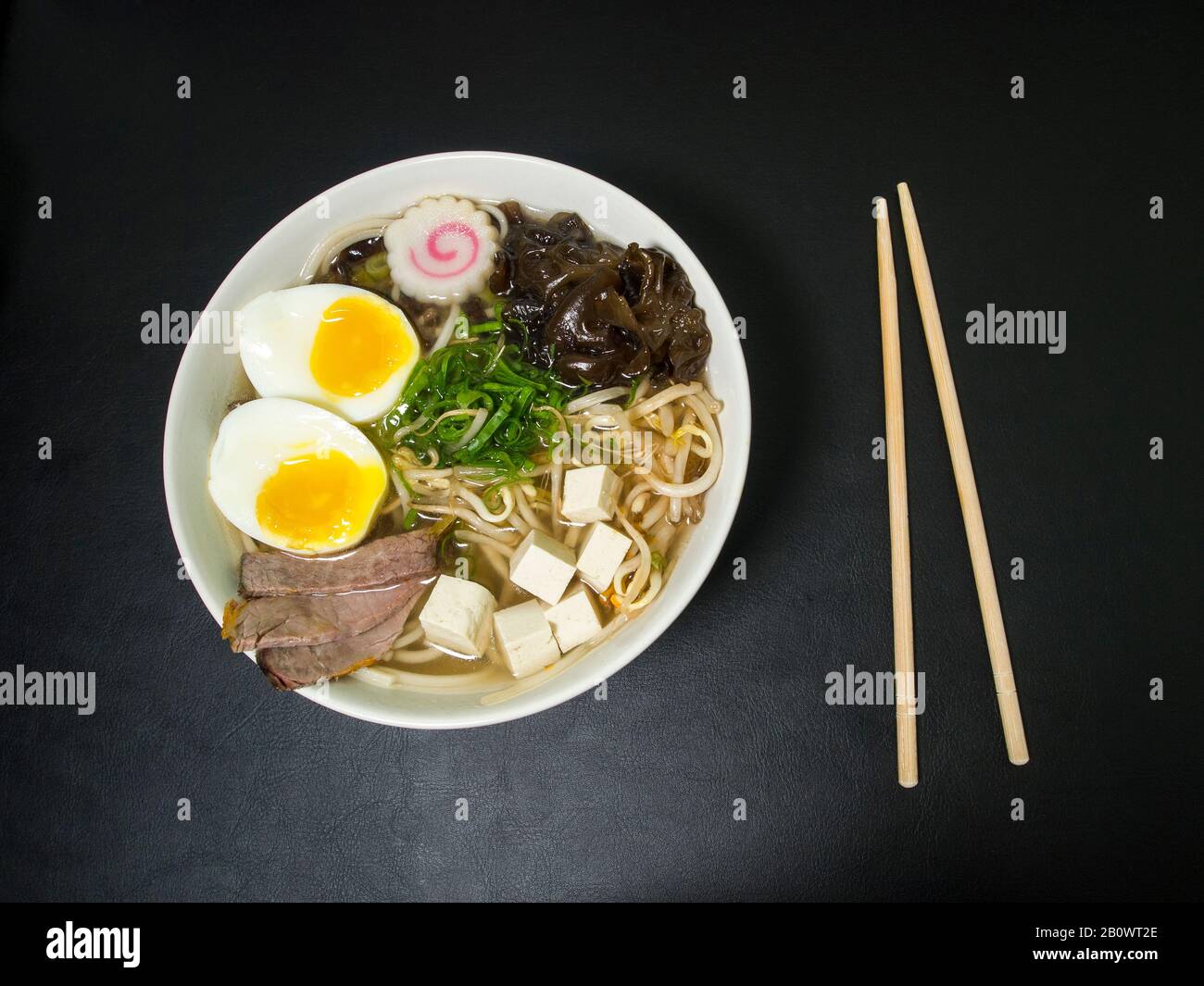 Ramen: Asian noodle soup with beef, eggs, fungi, vegetables, tofu, seaweed and naruto in a bowl with chopsticks on the side. Black background. Top vie Stock Photo