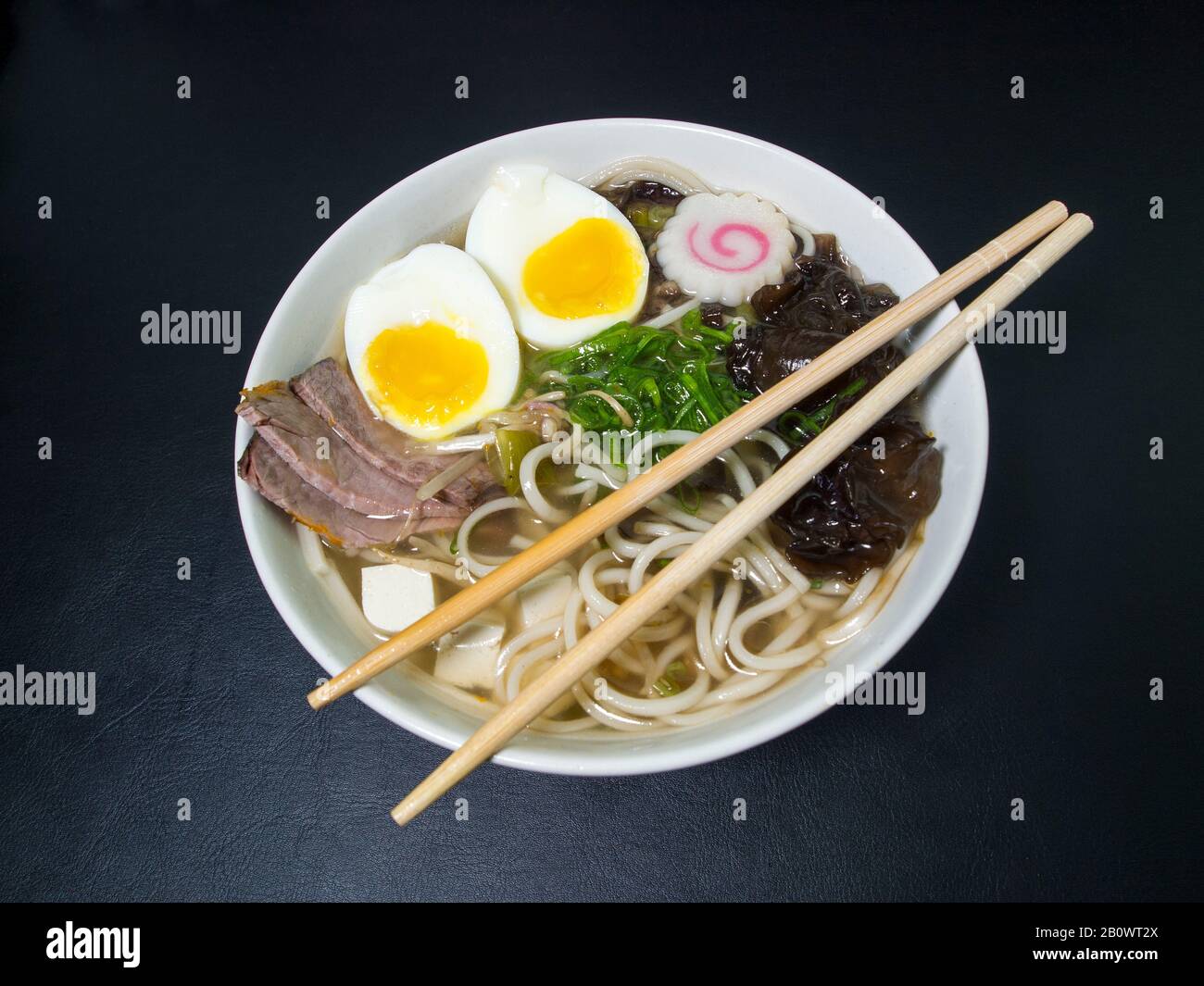 Ramen: Asian noodle soup with beef, eggs, fungi, vegetables, tofu, seaweed and naruto with chopsticks on the bowl. Black background. Top view. Stock Photo