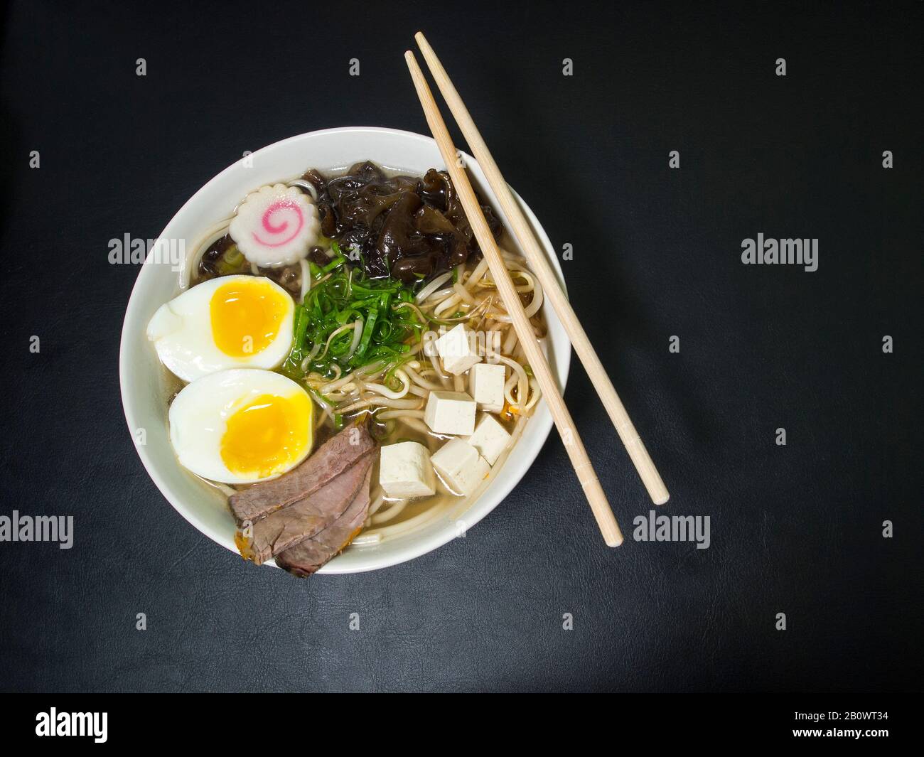 Ramen: Asian noodle soup with beef, eggs, fungi, vegetables, tofu, seaweed and naruto in a bowl with chopsticks. Black background. Top view. Stock Photo