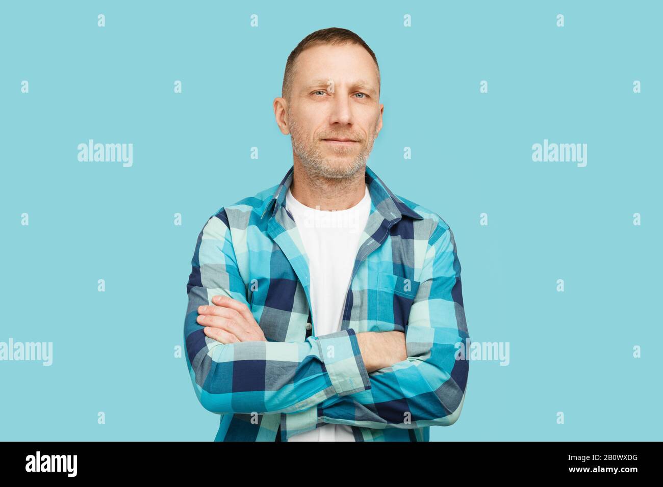Bearded man crossed arms wearing a white t-shirt and blue checkered shirt on a blue background. Stock Photo