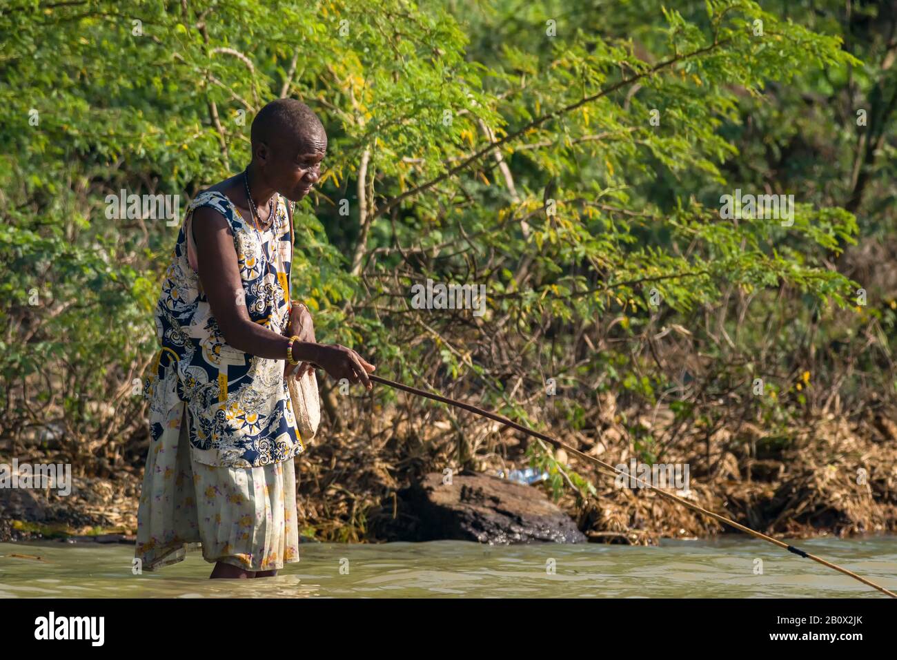 An elderly female Chamus or Njemps Kenyan woman standing in lake Baringo fishing by shore with fishing rod, Kenya Stock Photo