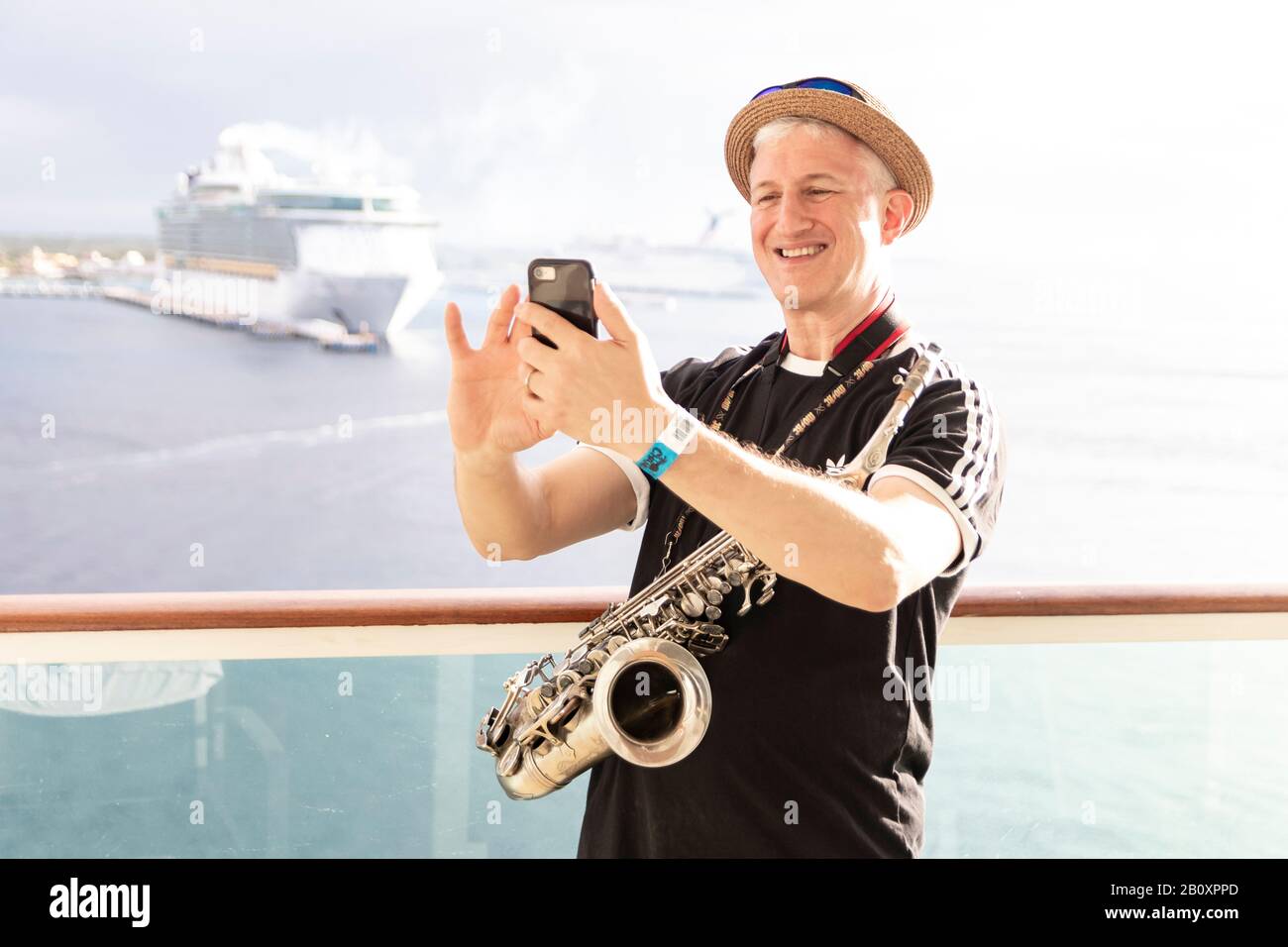 Jolly good time in the Caribbean sun; a smiling saxophone man takes a cell phone photo with a cruise ship at sea in the background. Stock Photo