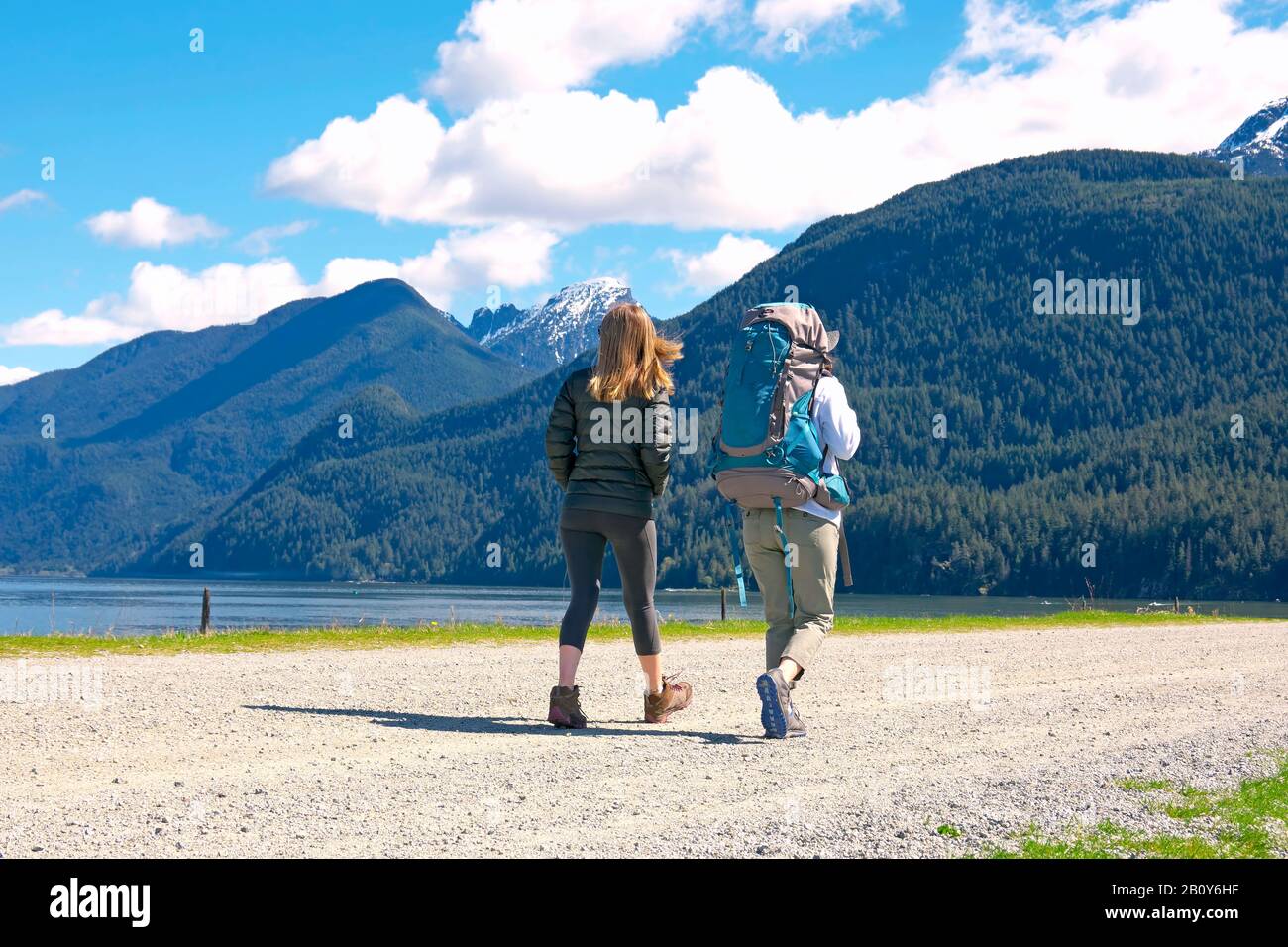 Two young women walking on gravel road beside a lake with mountains in the background.  Stock photo. Stock Photo