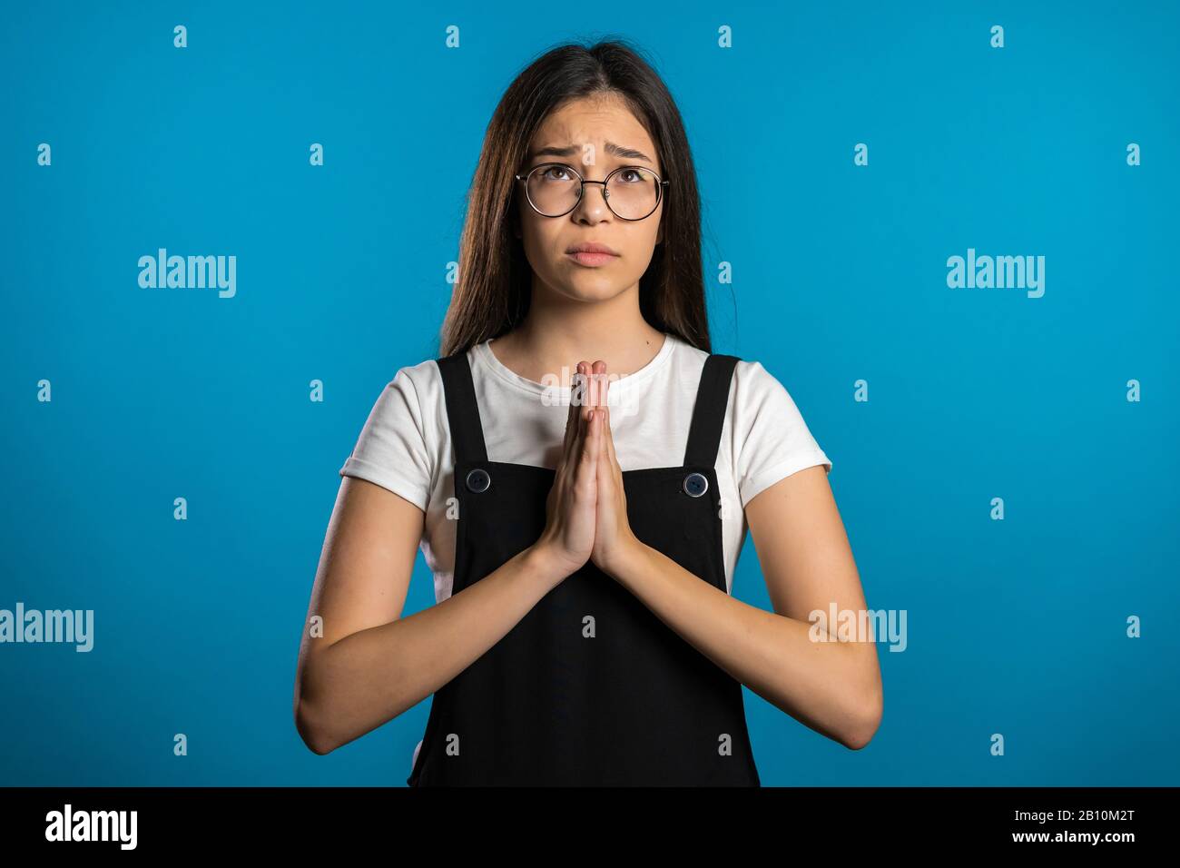 Cute asian young girl praying over blue background. Woman in glasses begging someone.  Stock Photo