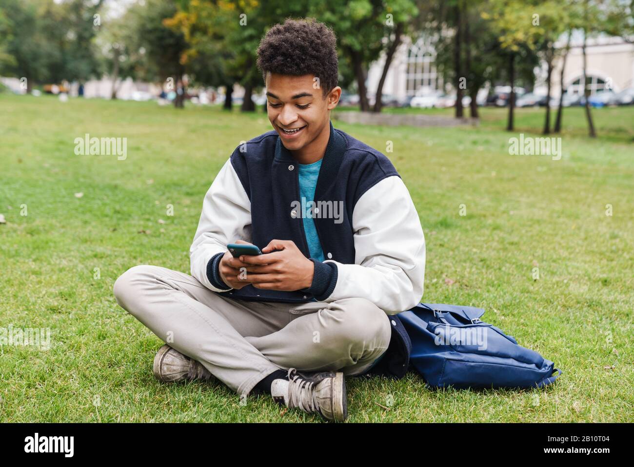 Happy teenager student boy sitting on a lawn at the park, using mobile phone Stock Photo