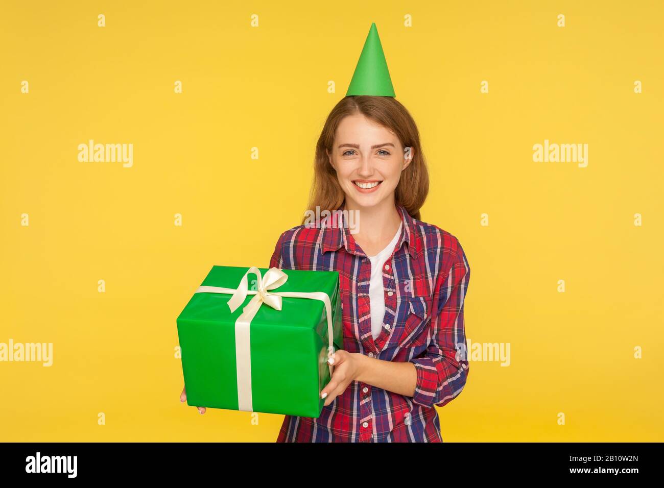 Portrait of cheerful funny ginger girl with party cone on head holding gift box and smiling happily, excited about present on birthday, holiday bonus. Stock Photo