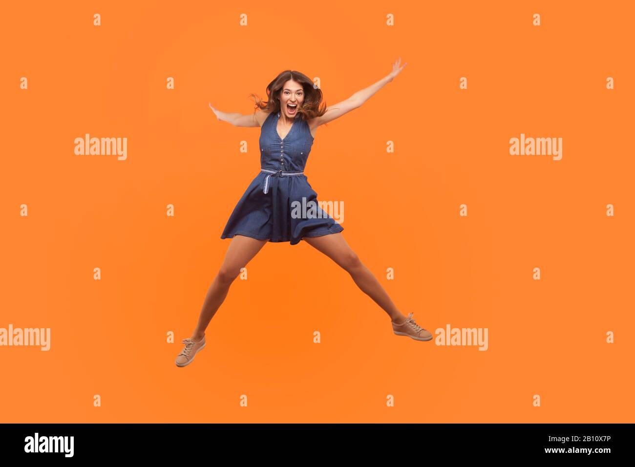 Overjoyed excited ecstatic brunette woman in denim dress jumping up like star and shouting from enthusiasm, flying isolated on orange background, full Stock Photo