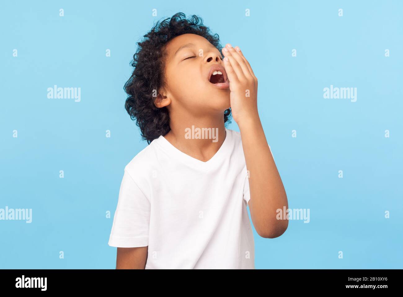Portrait of sleepy tired little boy with curly hair in white T-shirt covering mouth with hand while yawning with eyes closed, drowsy child waking up. Stock Photo