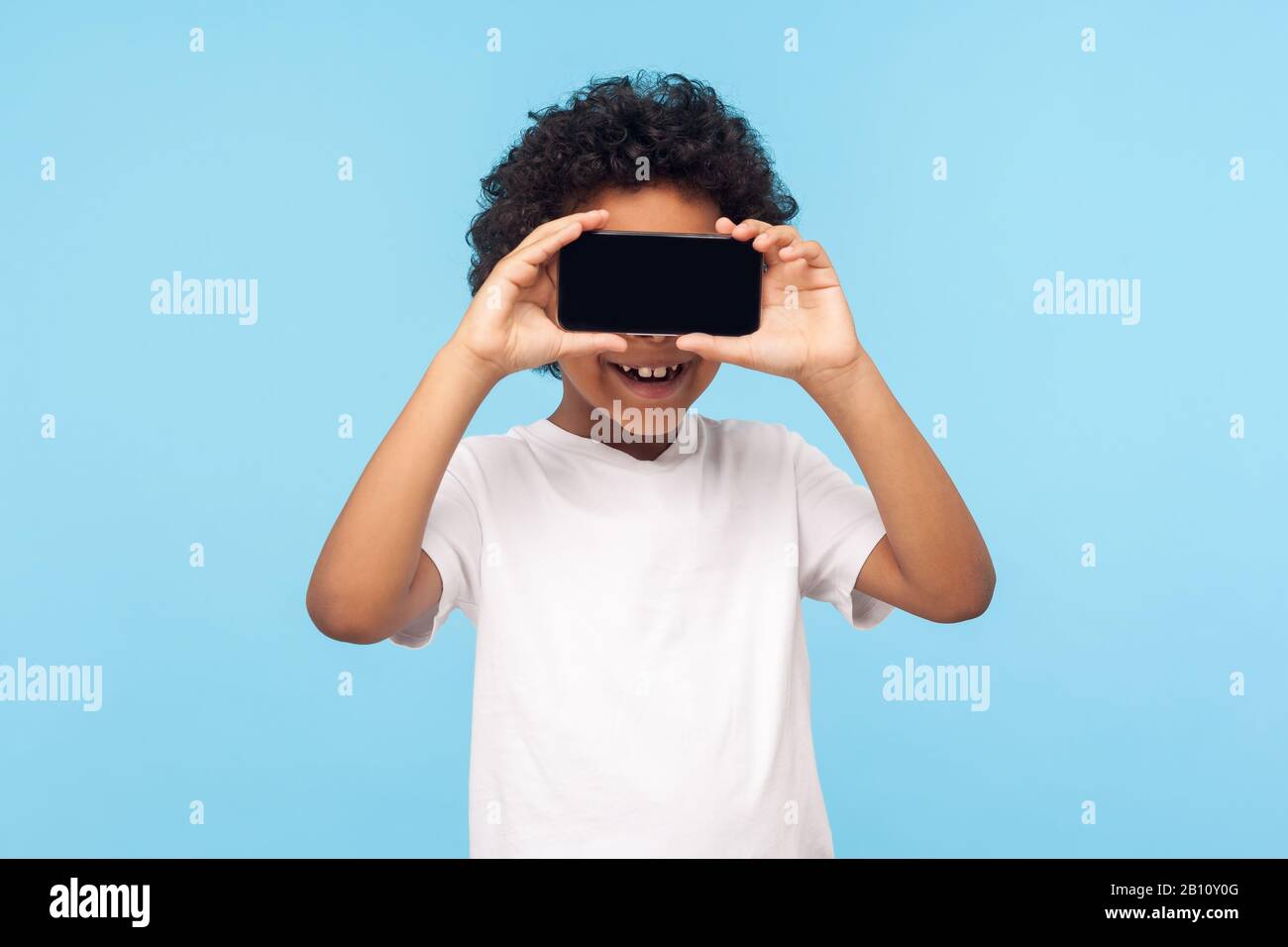 Positive lovely happy little boy with curly hair covering eyes with cellphone and smiling, unknown child hiding face with mobile phone, anonymous user Stock Photo