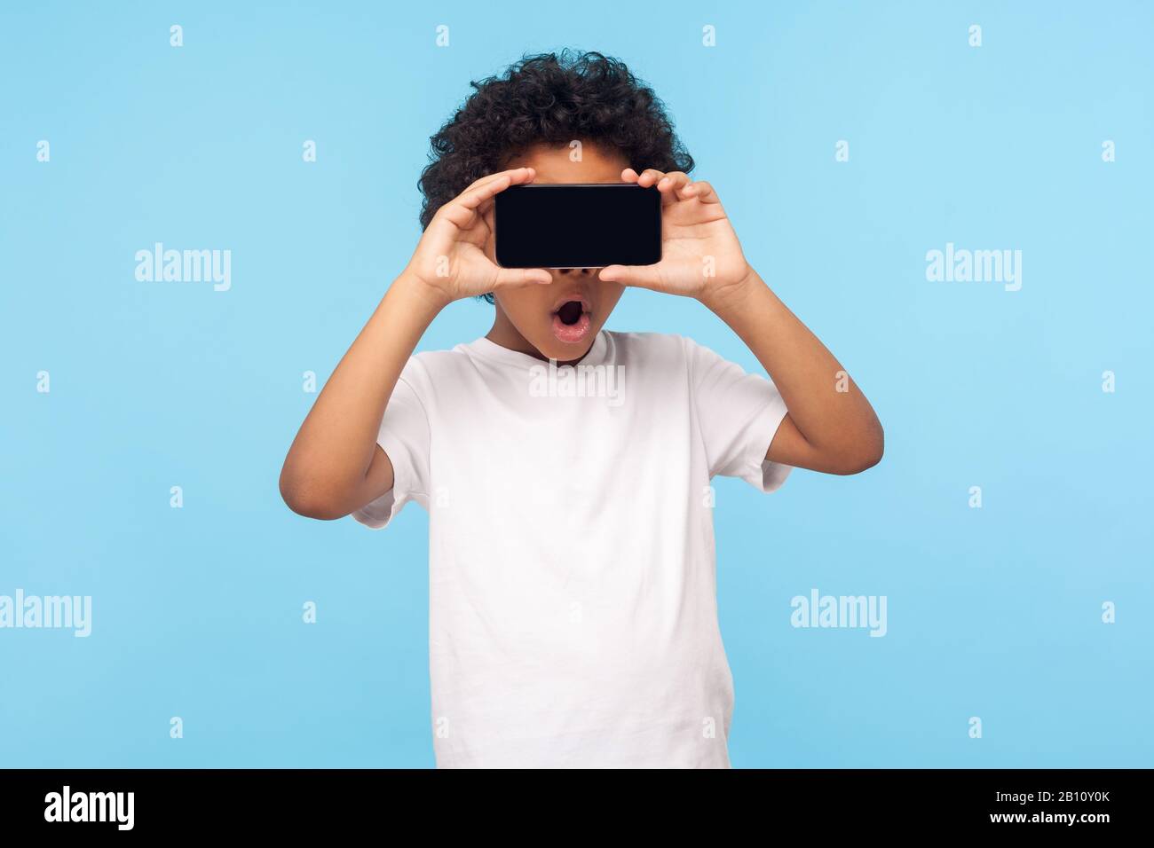 Funny amazed little mixed race boy covering eyes with cellphone, his mouth wide open from shock surprise, unknown child hiding face with mobile phone, Stock Photo