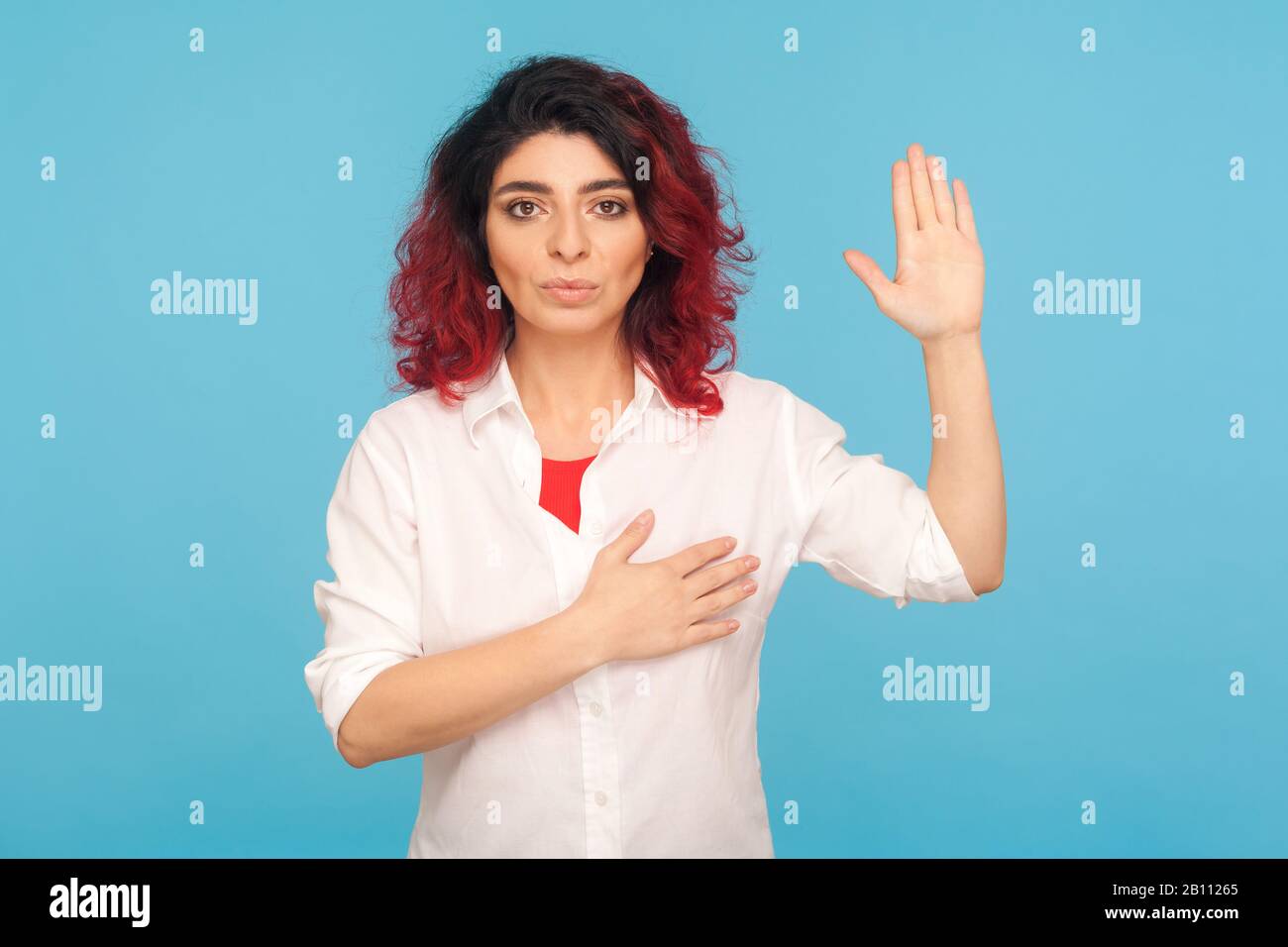 I swear! Portrait of hipster woman with fancy red hair holding hand on heart and raising palm to make oath, devoting herself, promising to fulfill tas Stock Photo