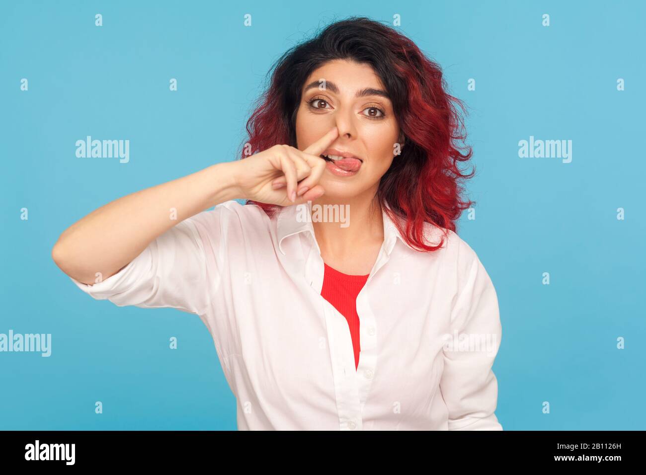 Bad manners, misconduct. Portrait of funny comical hipster woman with fancy red hair picking her nose and sticking out tongue with stupid expression. Stock Photo