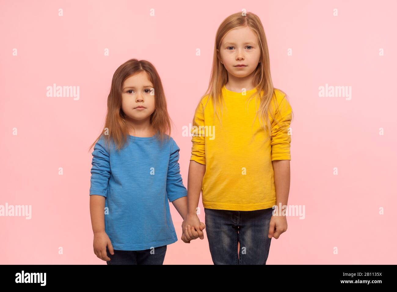 Little friends. Two cute charming preschool girls in sweatshirts standing together, holding hands and looking at camera with serious calm expression. Stock Photo
