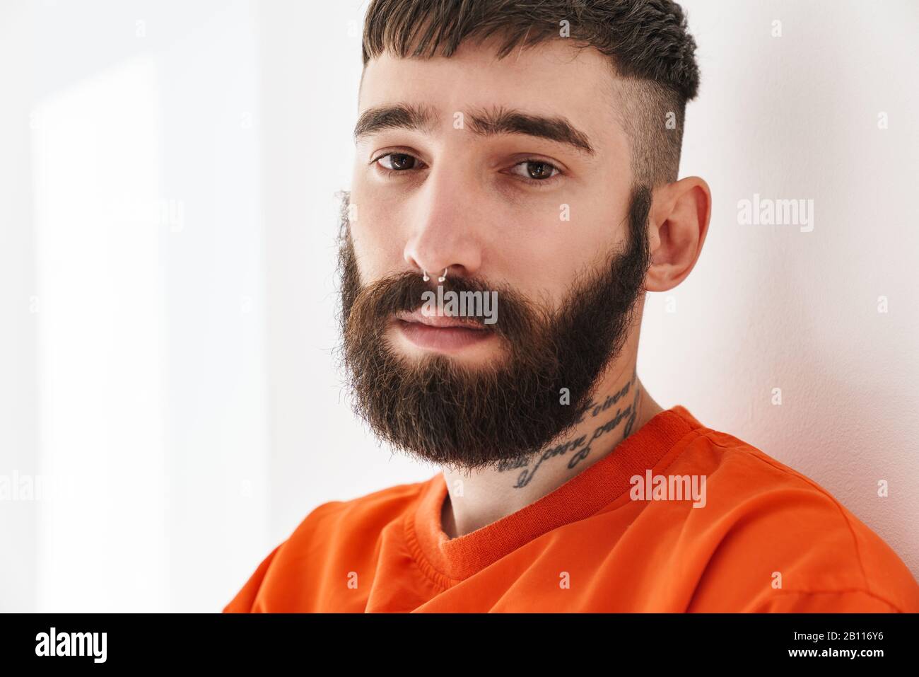 Image closeup of young bearded man with nose jewelry wearing orange shirt standing over white wall indoors Stock Photo