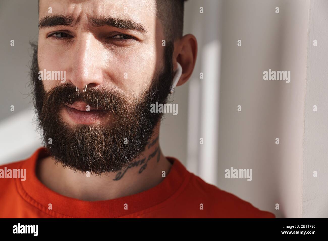 Image closeup of young bearded man with nose jewelry wearing earpods standing over white wall indoors Stock Photo