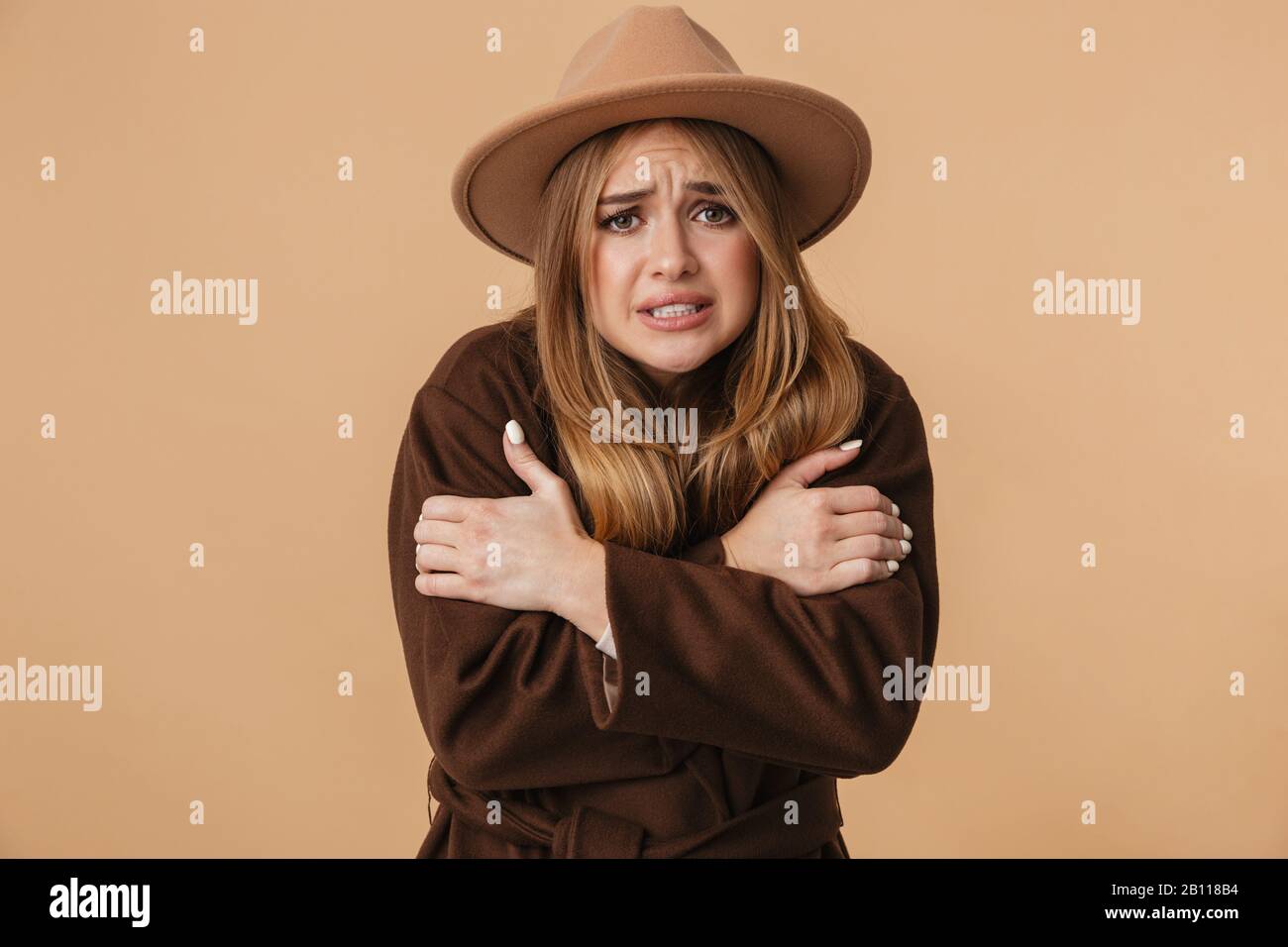 Image of young caucasian girl wearing hat and coat trembling and feeling cold isolated over beige background Stock Photo
