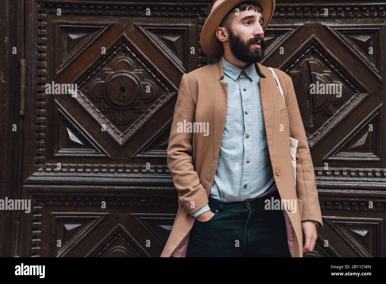 Attractive young man wearing autumn coat and hat standing outdoors at the city street in front of the wooden door Stock Photo