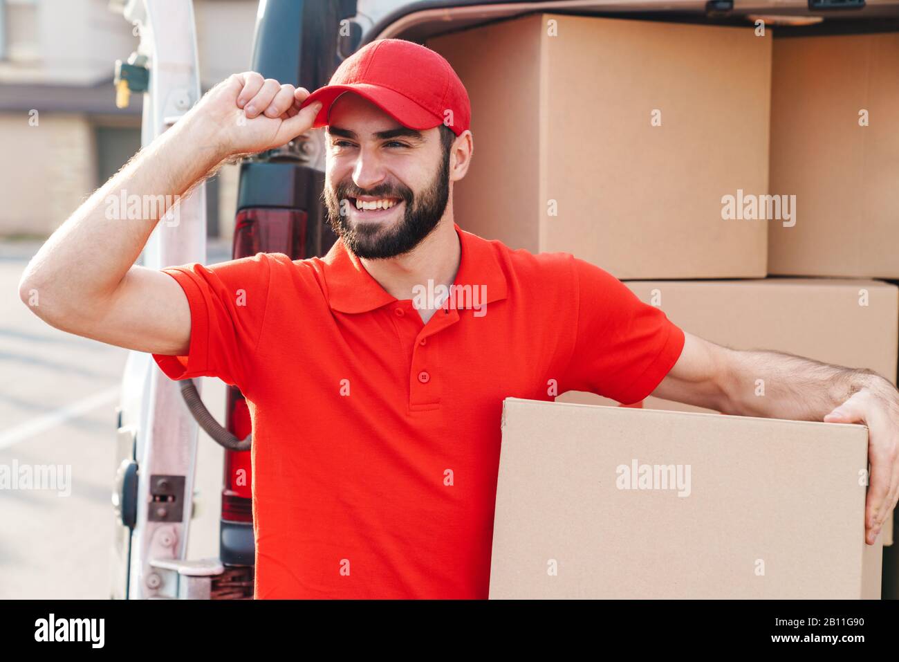Image of happy young delivery man in red uniform standing with parcel ...