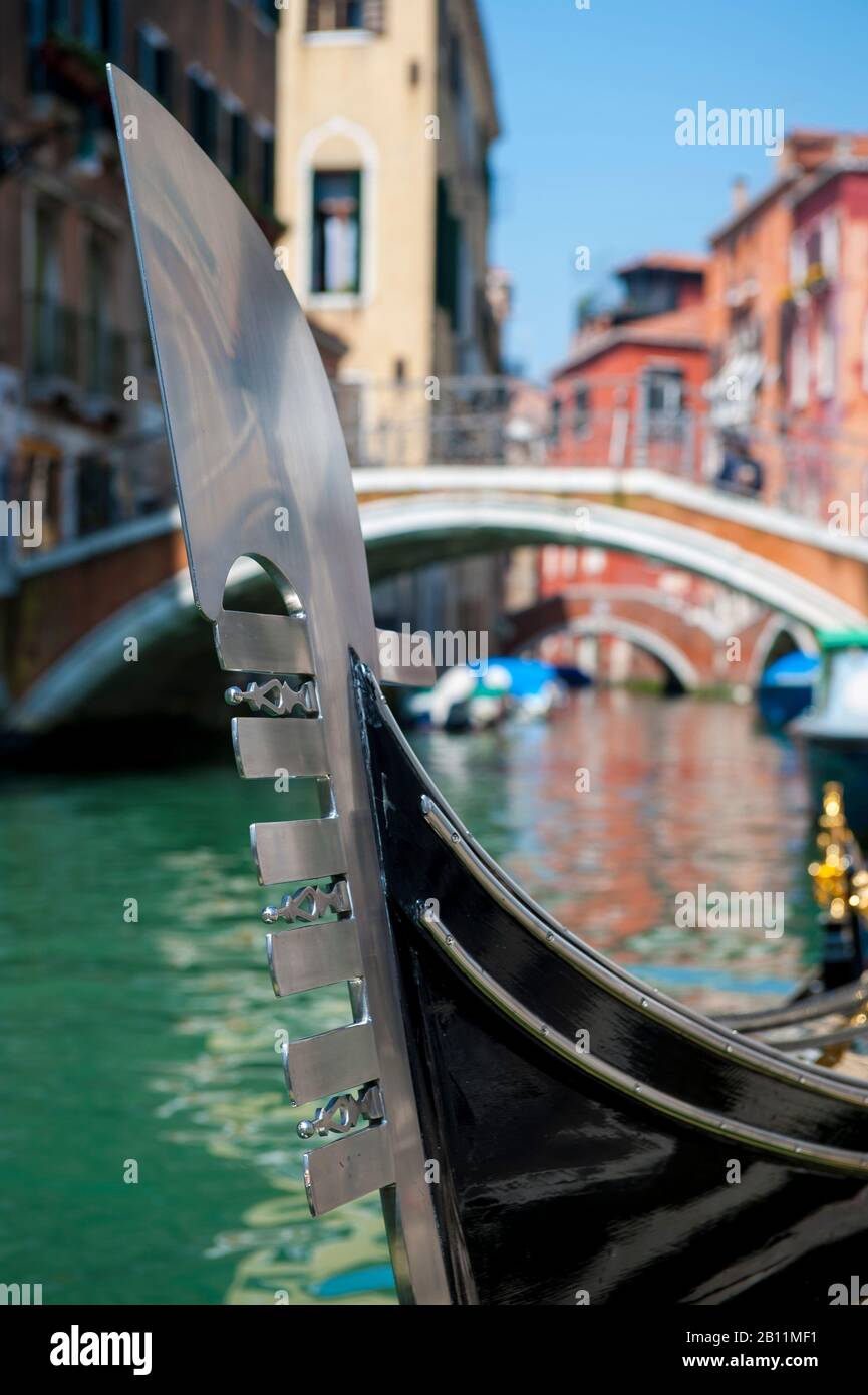 Close-up of a traditional "ferro" metal bow on a Venetian gondola in front of a bright sunny canal view of Venice, Italy Stock Photo