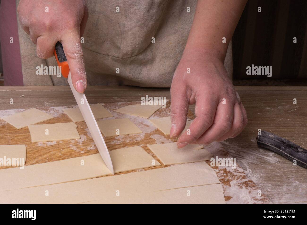 A woman cuts the dough into squares with a white ceramic knife. Plywood cutting board, wooden flour sieve and wooden rolling pin - tools for making Stock Photo