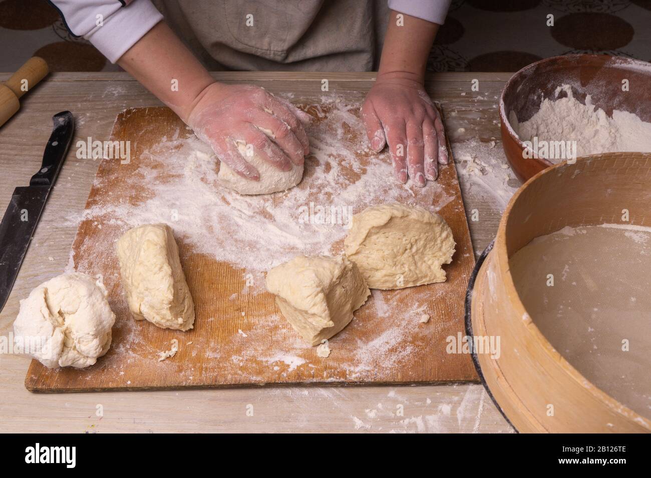 A woman kneads the dough. Plywood cutting board, wooden flour sieve and wooden rolling pin - tools for making dough. Stock Photo