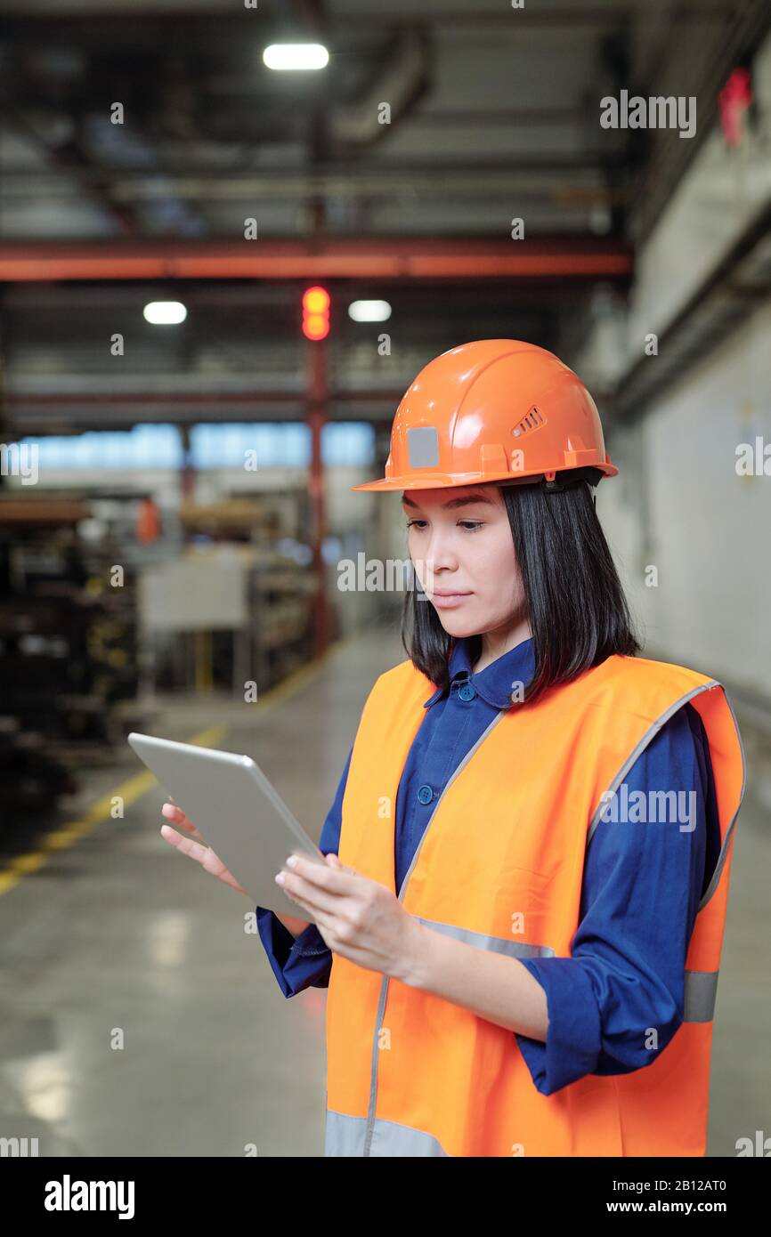 Pretty young serious female engineer in hardhat and workwear using touchpad Stock Photo