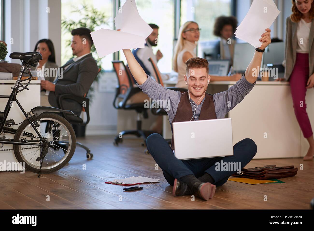 young man at the office sitting o n the floor with his laptop with hands holding papers in the air after receiving good business news. casual, busines Stock Photo