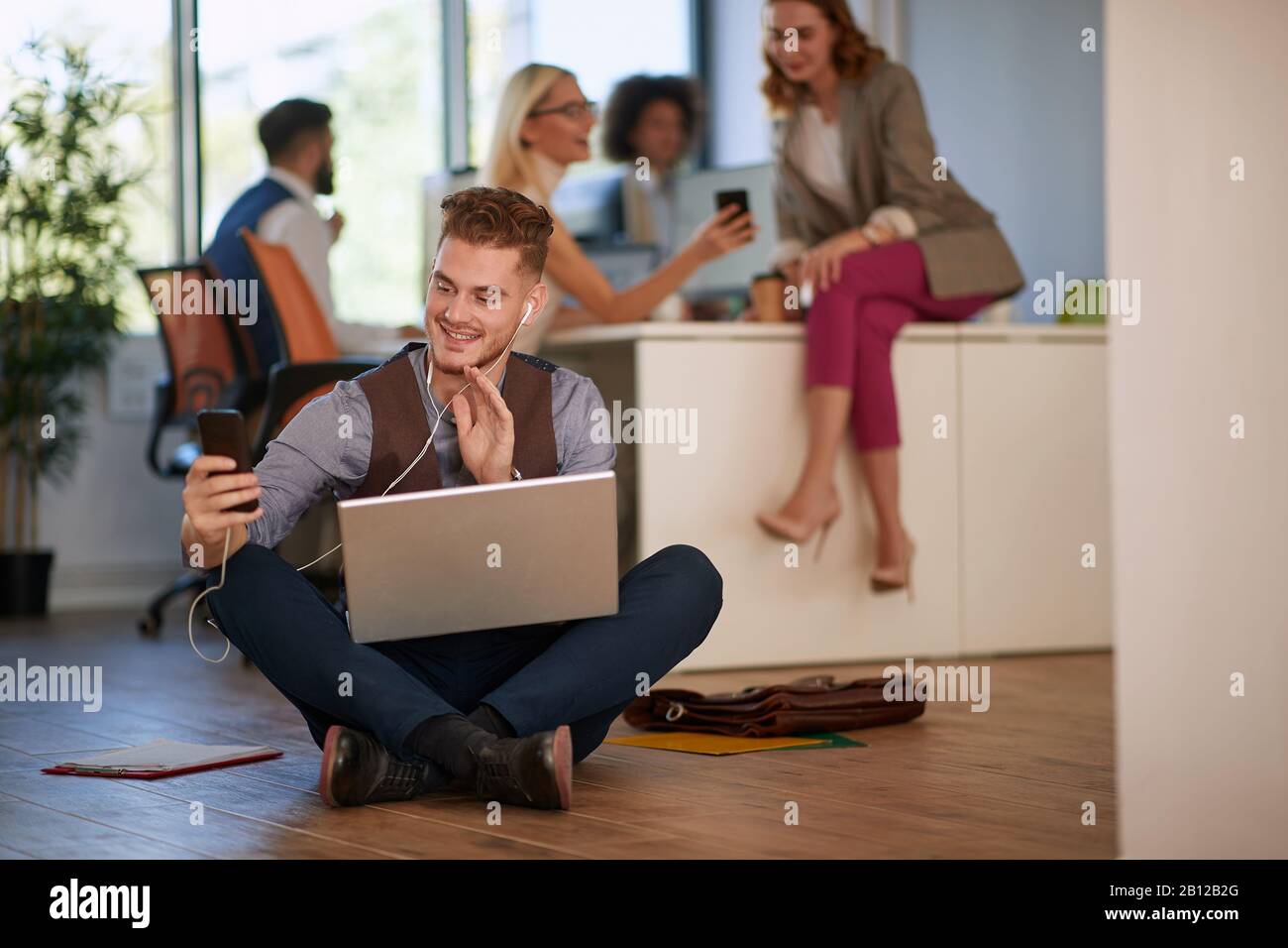 man at the office sitting on the floor with his laptop using his cell phone. casual, business, concept Stock Photo