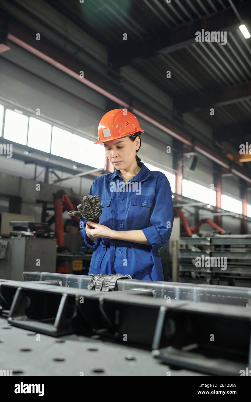 Young serious female engineer in workwear putting on gloves before work
