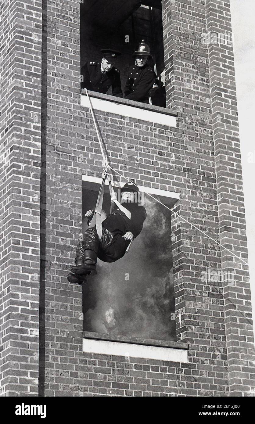 1970s, historical, a fireman in a harness and pulley escaping from a smoke-filled brick-built tower during a training drill or exercise, watched from above by senior fire officers, England, UK. Stock Photo