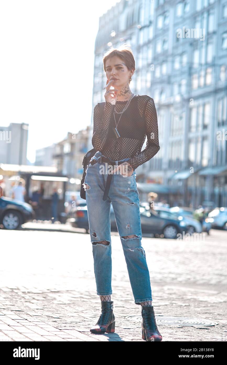 Urban Style. Young stylish woman standing hand in pocket outdoors smoking  cigarette posing cool Stock Photo