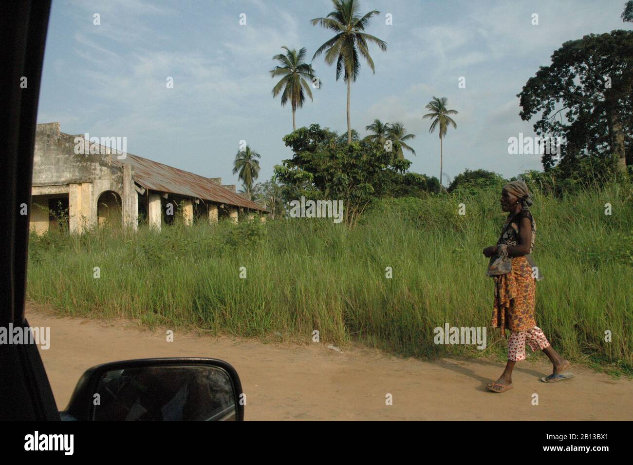 Walking African woman on street of sand with high green meadow, palms and an African church in background. wearing brown colored clothes with African Stock Photo