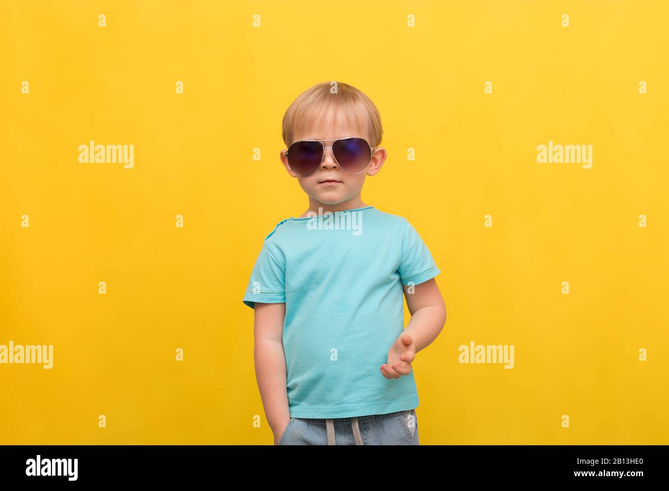 Child portrait of a 3 years old blond boy in a blue T-shirt and sunglasses on a bright yellow spring summer background with place for text. Attracting Stock Photo
