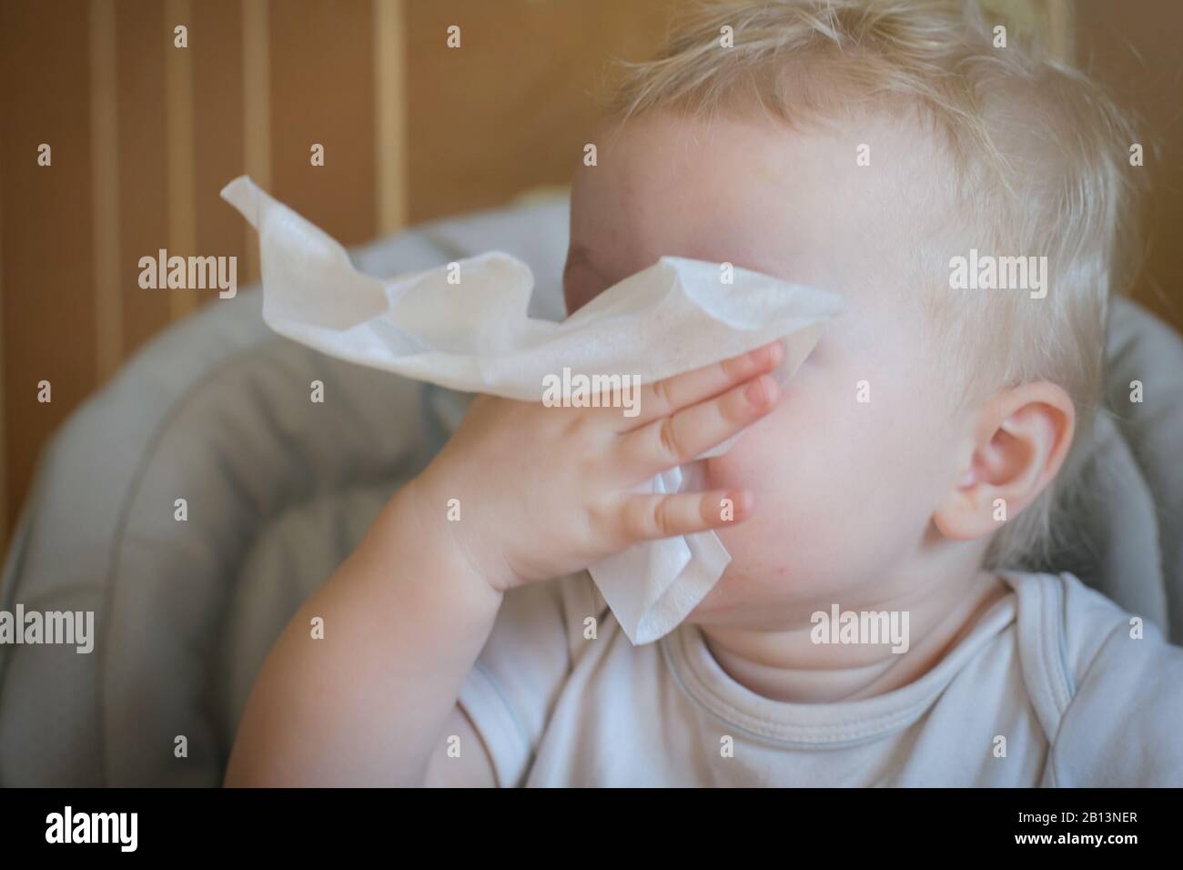 The child wipes his nose with a white handkerchief. A sick child eats sitting on feeding chair. Kid with cold rhinitis. virus and infection. Coronavir Stock Photo