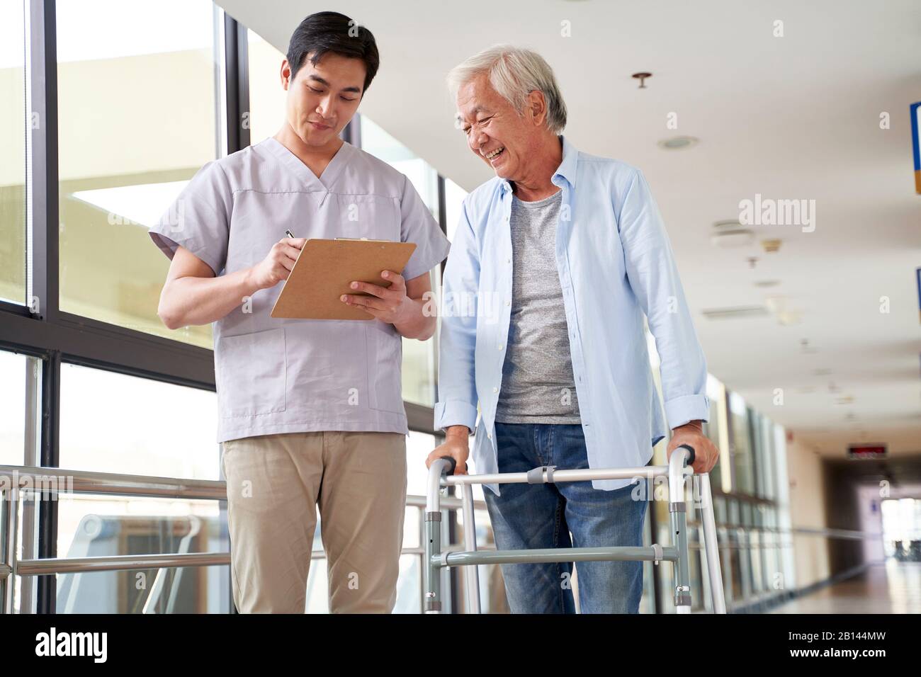 young asian physical therapist working with old man on working using a walker in hallway of nursing home Stock Photo