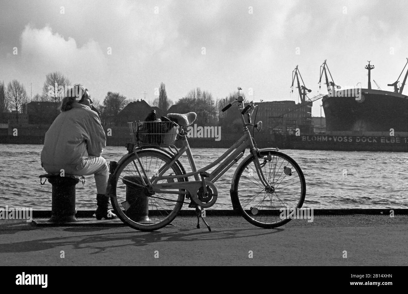 Lady sits on a bollard with her bicycle on the quayside on St. Pauli Landungsbrücken, looking over the Elbe to the shipbuilding complex of Blohm & Voss, Hamburg, Germany, circa 1988.  Black and white film photograph Stock Photo