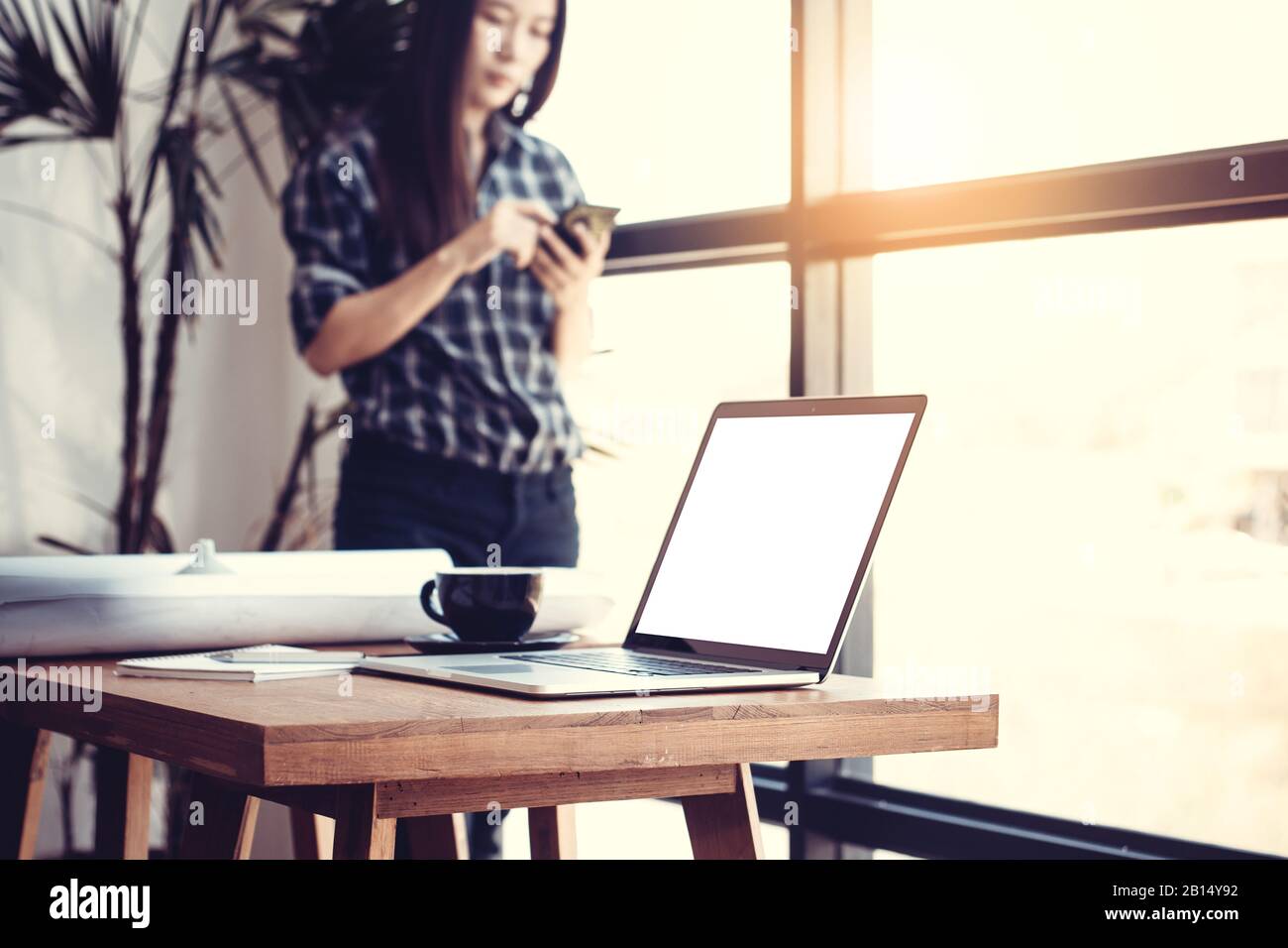 Technology in workplace concept. Young Asian business woman in modern office using the phone for work. Blank white screen laptop and blueprint on desk Stock Photo