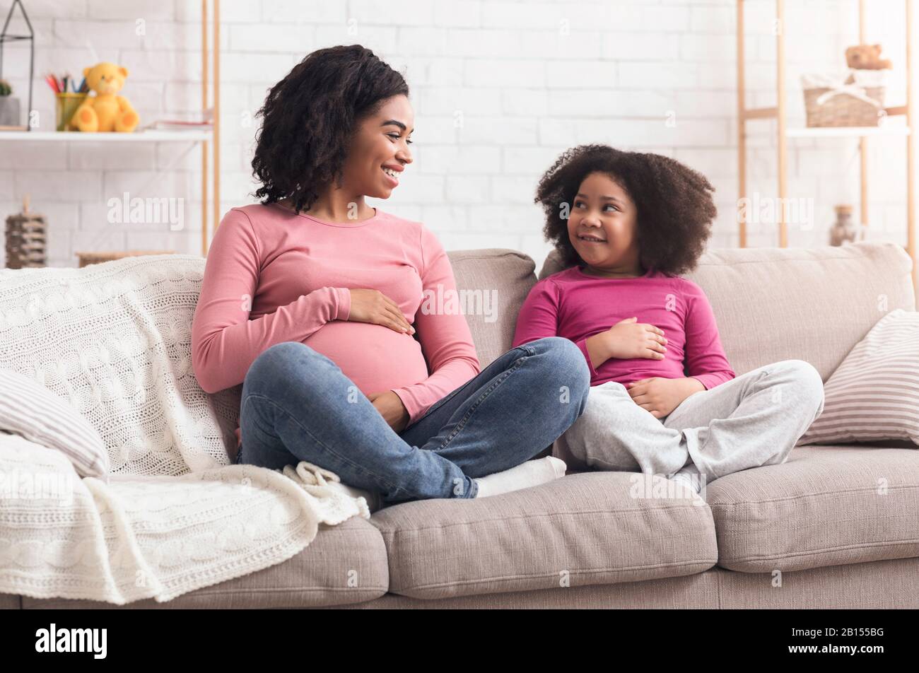 Pregnant afro mom and her daughter touching bellies, sitting on couch