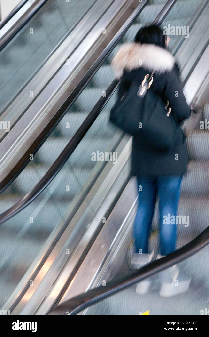 Young teenager woman in movement standing alone in an urban up moving escalator leaving  the subway station traveling upstairs to the city mall. Stock Photo