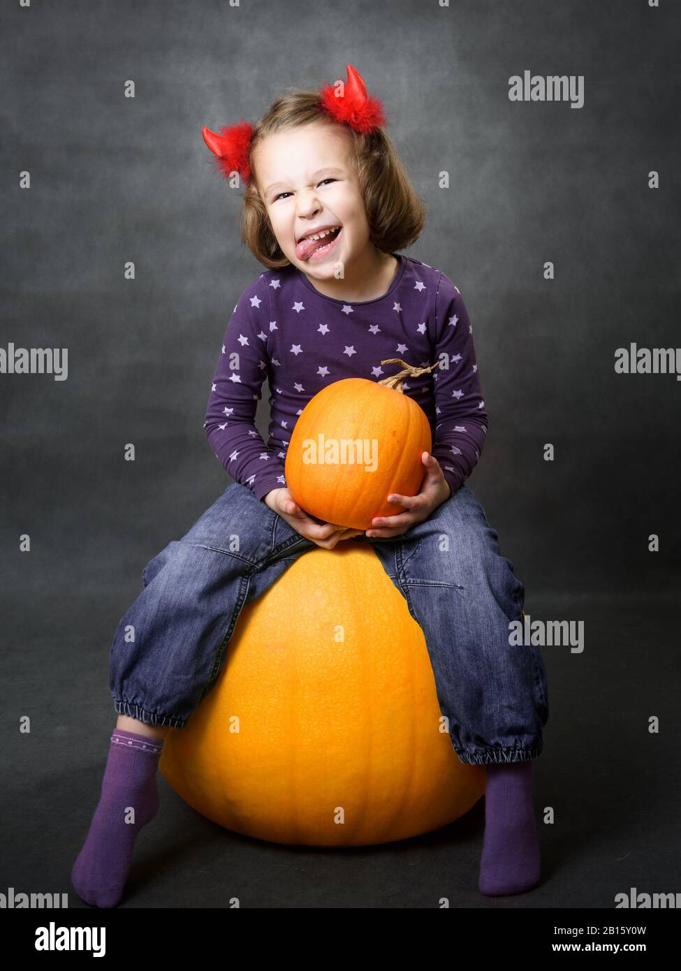 Halloween concept. Little girl with costume horns having fun. Adorable child sits on pumpkin and grimaces. Toddler smiles and shows tongue. Studio por Stock Photo