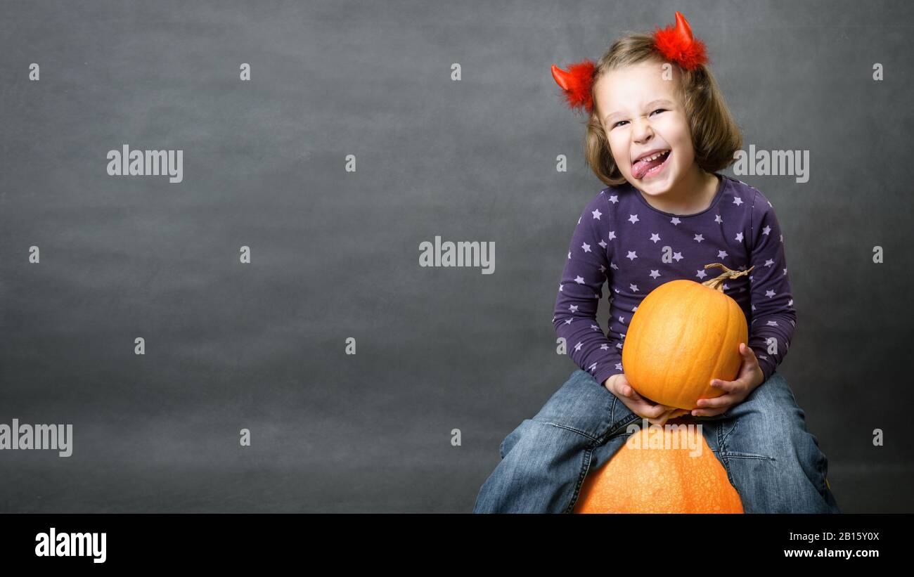 Halloween background with adorable child and space for your text. Little girl with costume horns having fun. Cute toddler sits on pumpkin and shows to Stock Photo