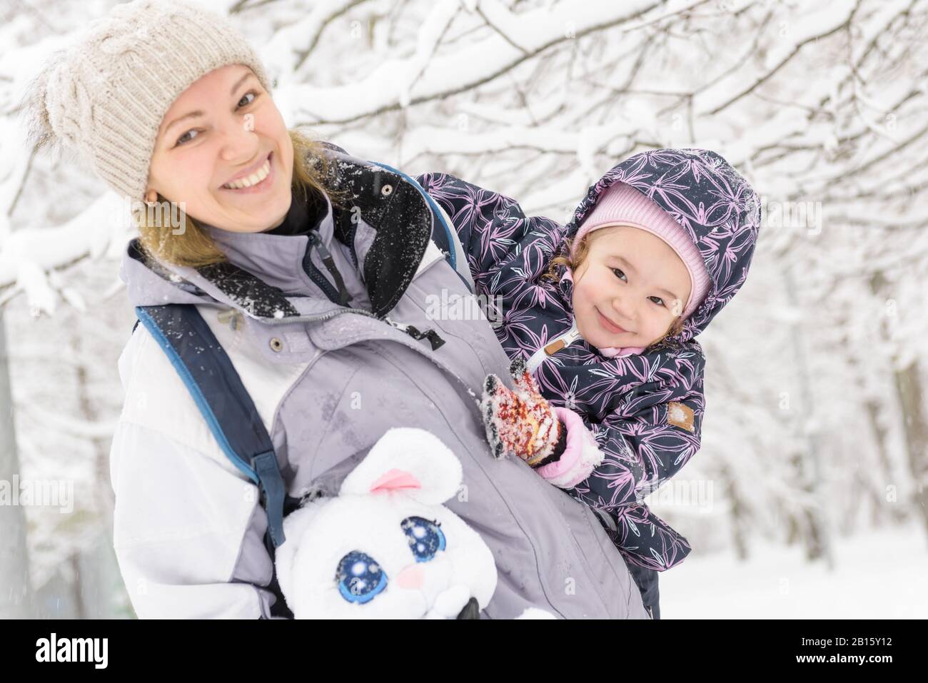 Cute cheerful little child plays with mother in a snowy park in winter. Happy family walks during snowfall. Stock Photo