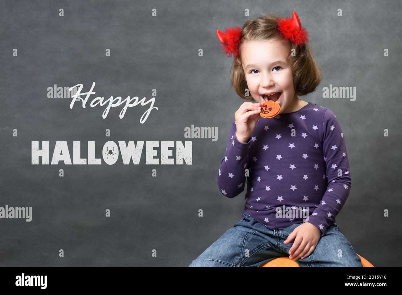 Happy Halloween! Little girl with costume horns having fun. Adorable child with Halloween typography. Cute toddler smiles and eats Halloween treats of Stock Photo