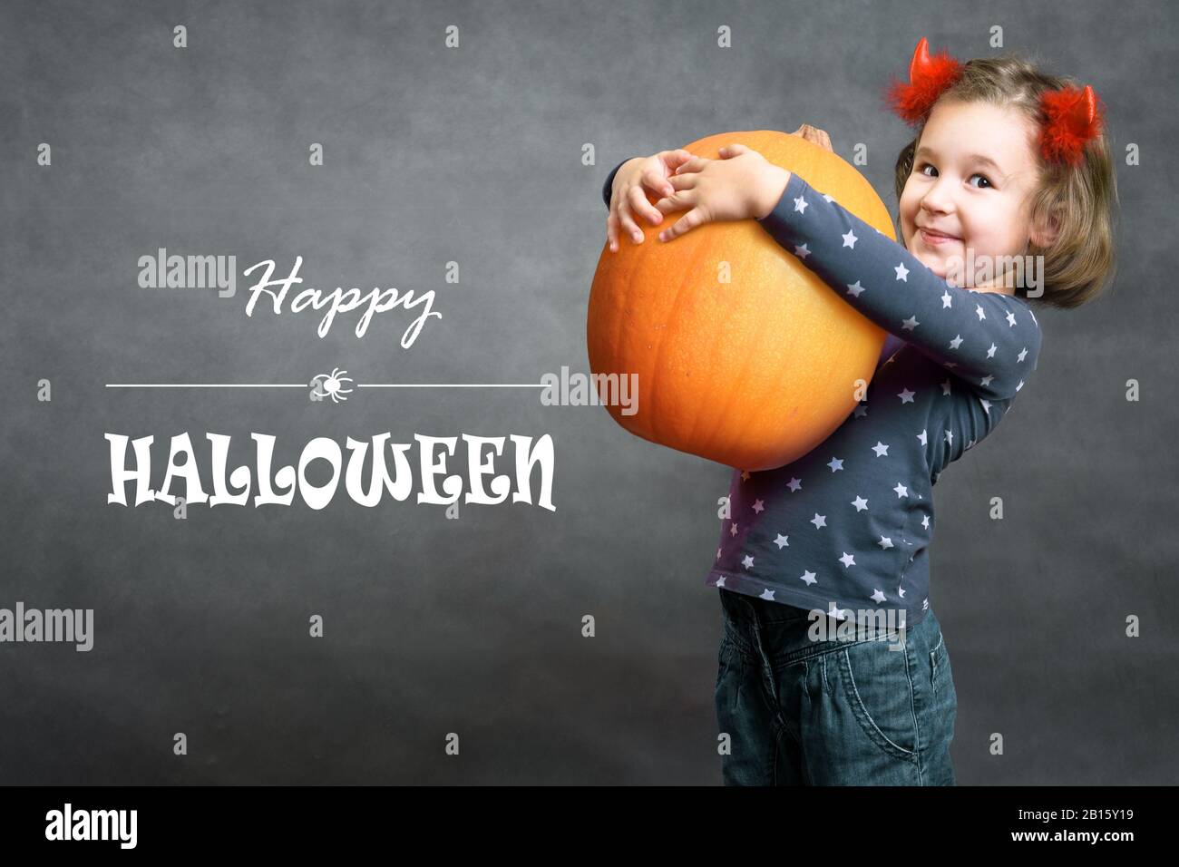 Happy Halloween! Funny little girl holds big pumpkin and smiles. Adorable child and Halloween typography. Cute toddler with costume horns having fun. Stock Photo