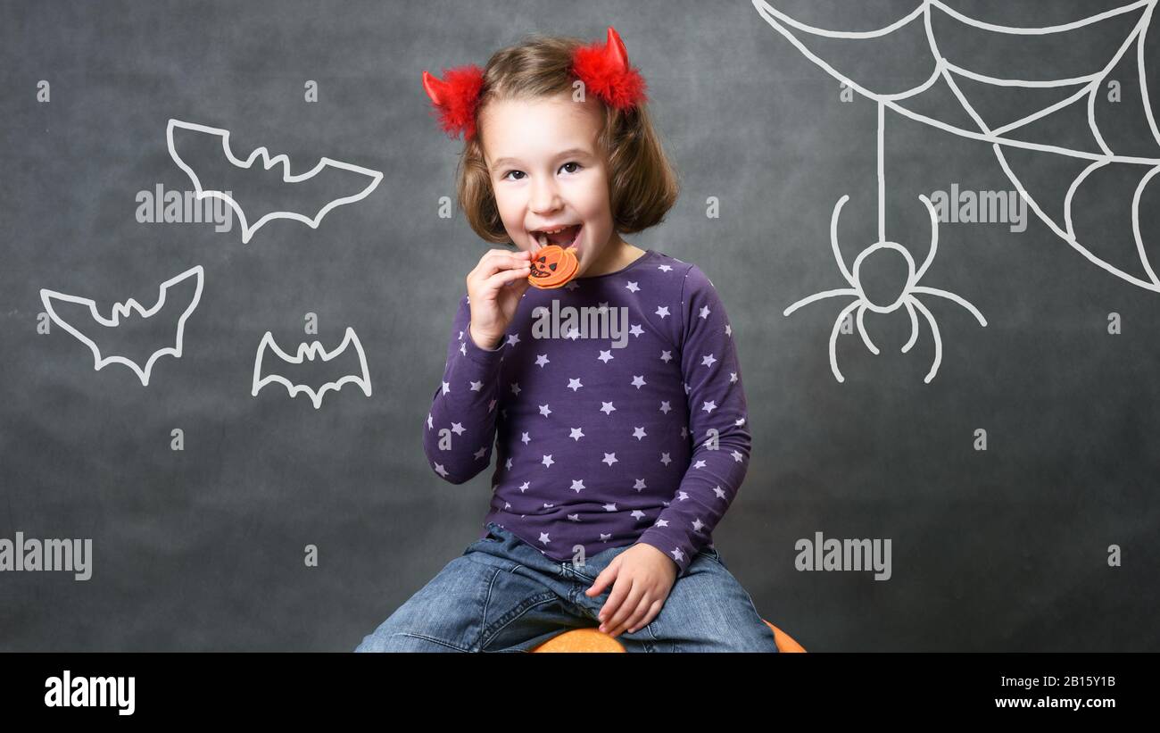 Halloween concept. Little girl with costume horns having fun. Adorable child with bats and spider. Cute toddler smiles and eats Halloween treats of pu Stock Photo