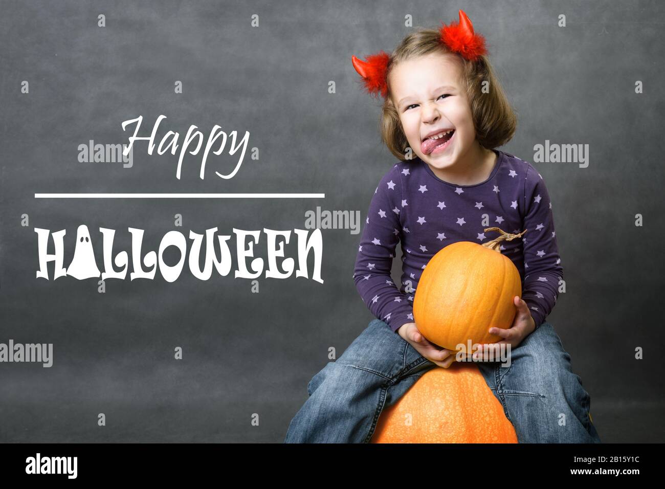 Happy Halloween! Little girl with costume horns having fun. Adorable child with pumpkin and Halloween typography. Cute toddler smiles and shows tongue Stock Photo