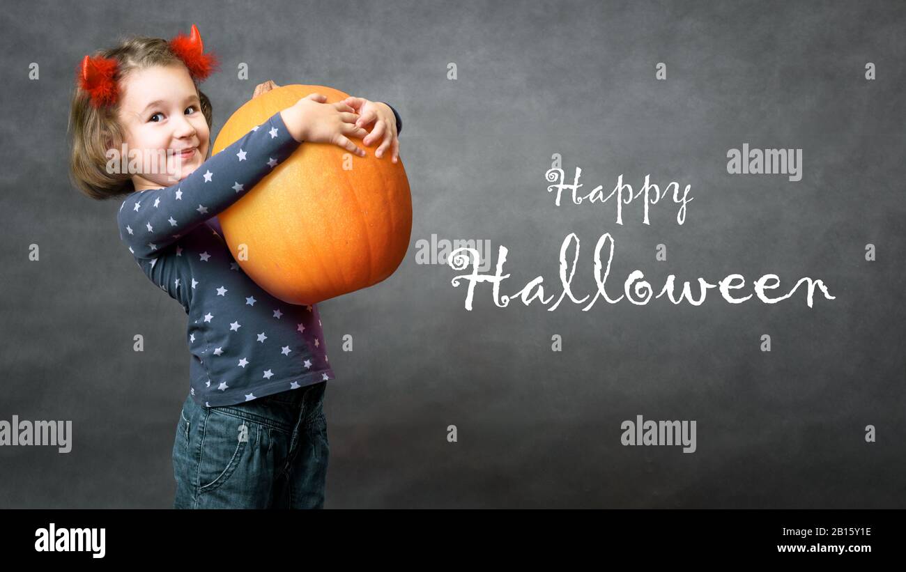 Happy Halloween! Little girl with costume horns having fun. Adorable child and Halloween typography. Cute funny toddler holds big pumpkin and smiles. Stock Photo