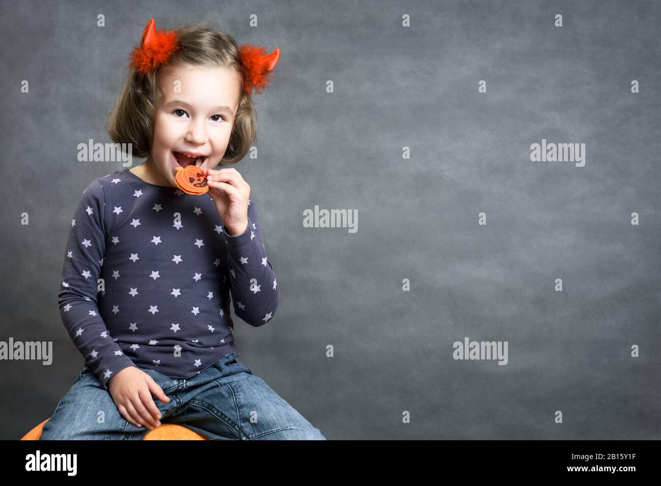 Halloween background with adorable child and space for your text. Little girl with costume horns having fun. Cute toddler smiles and eats Halloween tr Stock Photo