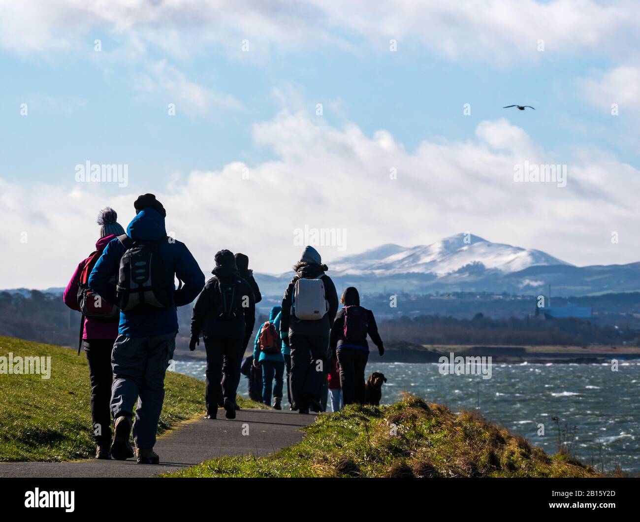 Firth of Forth, East Lothian, Scotland, United Kingdom, 23rd February 2020. UK Weather: Very strong wind on the coastline with sunshine. The wind creates white caps in the sea and big waves against the shore. A group of walkers on the John Muir Way with snow on the Pentland Hills in the distance Stock Photo