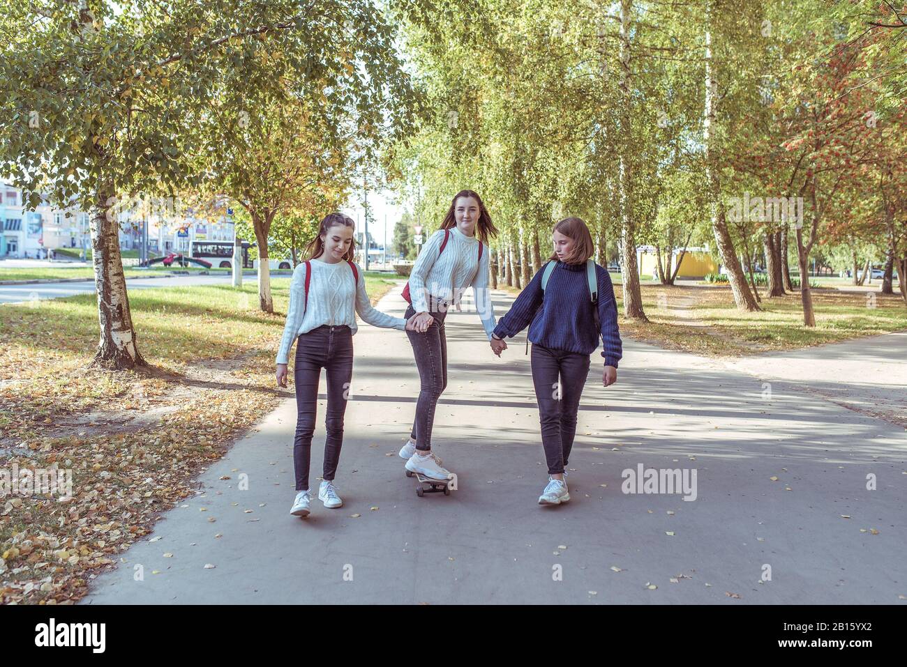 Three schoolgirls girls girlfriends in fall in city, ride a skateboard, in summer in park, return from school and college, behind back of bag are Stock Photo